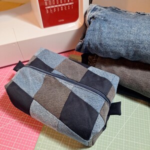 Peut inclure: Une trousse à crayons en patchwork de denim avec une fermeture éclair et des tirettes bleu marine. L'étui est fait de carrés de denim bleu, gris et noir. Une machine à coudre et des jeans en denim pliés sont en arrière-plan.