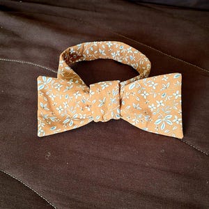 Peut inclure: Un nœud papillon marron avec un motif floral blanc. Le nœud papillon est en tissu et a une forme classique.