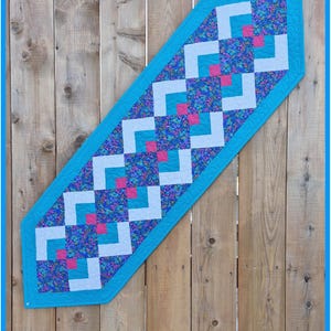 May include: A table runner with a blue border and a geometric pattern of white, blue floral, and pink squares. The text "Wild Daisy Quilts" and "Junior Jiffy Runner" are visible.