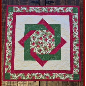 Puede incluir: Un adorno cuadrado acolchado con un diseño floral y geométrico. El centro presenta un estampado floral rodeado de cuadrados entrelazados verdes y rojos. El borde tiene un estampado floral sobre un fondo crema. El texto "Wild Daisy Quilts" y "Interlocking Squares Topper" son visibles.