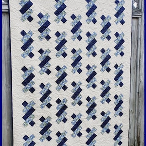 Peut inclure: Une courtepointe avec un motif "Twisties" en tissu bleu et blanc. La courtepointe est composée de carrés avec un motif diagonal. La courtepointe est exposée contre une clôture en bois.
