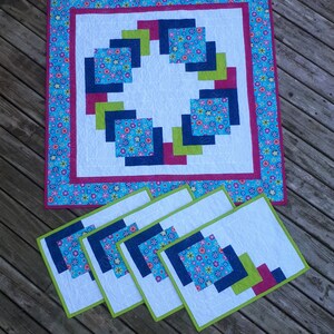 Pu&ograve; includere: Set di cinque tovagliette all'americana con un design geometrico che presenta quadrati in tonalit&agrave; di blu, verde e rosa. Le tovagliette hanno uno sfondo bianco e sono bordate di verde.