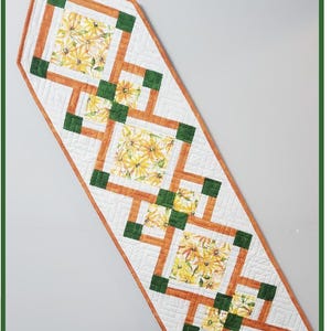 Pode incluir: Um corredor de mesa com um padrão geométrico de quadrados laranja e verde. Os quadrados são preenchidos com uma estampa floral de margaridas amarelas sobre um fundo branco. O texto "The Everyday Runner" está na parte inferior da imagem.