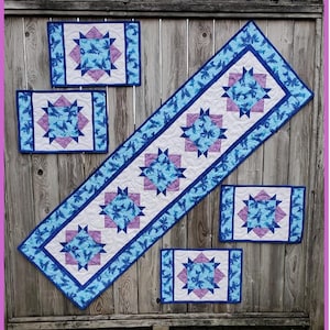 Pode incluir: Um conjunto de quatro tapetes individuais acolchoados azuis e roxos e um corredor de mesa correspondente. Os tapetes individuais e o corredor de mesa apresentam um design de estrela com fundo branco. O texto "EZ Place Mats & Runner" é exibido na parte inferior da imagem.