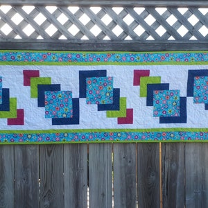 Pu&ograve; includere: Un runner da tavolo colorato con un motivo geometrico di quadrati in tonalit&agrave; di blu, verde e rosso. Il runner &egrave; realizzato in tessuto con stampa floreale e ha uno sfondo bianco.