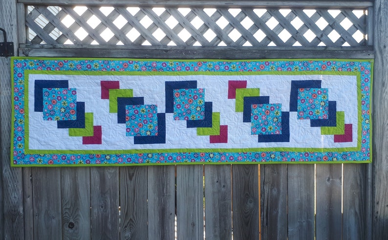 Pu&ograve; includere: Un runner da tavolo colorato con un motivo geometrico di quadrati in tonalit&agrave; di blu, verde e rosso. Il runner &egrave; realizzato in tessuto con stampa floreale e ha uno sfondo bianco.