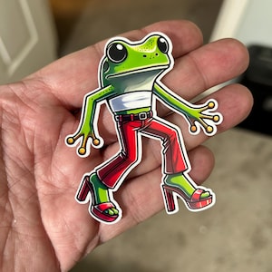 May include: A green frog wearing red pants and high heels. The frog is walking with its arms outstretched.