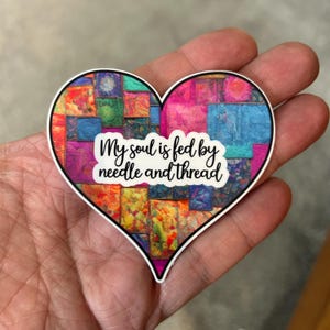May include: A heart-shaped sticker with a colorful patchwork design and the text "My soul is fed by needle and thread".