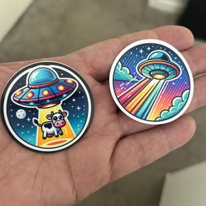 May include: Two round stickers featuring cartoon UFO designs. One shows a UFO beaming a cow, the other a UFO with rainbow beams. Both have a blue, purple, and orange color scheme with a white border.