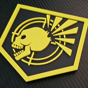 Könnte beinhalten: Ein gelbes Totenkopf-Emblem auf schwarzem Hintergrund, umrahmt von einem gelben rautenförmigen Rand. Der Schädel ist mit Zähnen und Augenhöhlen detailliert, mit stilisierten Sonnenstrahlen.