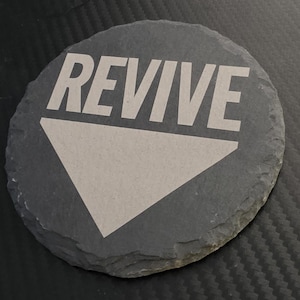 May include: A circular dark gray slate coaster with a rough, uneven edge. The word REVIVE is printed in a bold, light gray sans-serif font across the top half, positioned above a large, solid light gray downward-pointing triangle.