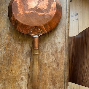 Op de afbeelding: Een koperen pan met een lange houten handgreep. De pan heeft een gehamerde textuur en een glanzend oppervlak. De handgreep is lichtbruin en heeft een lus aan het uiteinde. Een klassiek keukengerei.