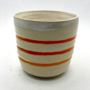 Op de afbeelding: Handgemaakte keramische mok met een crèmekleurige basis en horizontale banden in oranje en rood. De rand is een lichtere crèmekleur. De mok heeft een licht gestructureerd oppervlak en een afgeronde vorm.