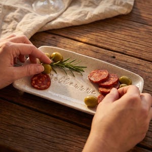 May include: A rectangular, speckled off-white ceramic serving tray featuring the embossed text "Melbourne" in the center. The tray is arranged with sliced red chorizo, several green olives, and a fresh sprig of green rosemary on its surface.