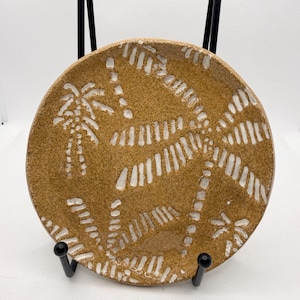 May include: A circular, tan ceramic plate featuring a raised white palm tree design. The plate is supported by a black metal stand. The palm tree design is repeated across the plate's surface.