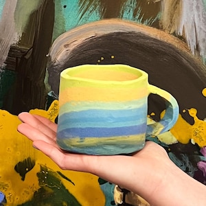 May include: A hand-held, ceramic mug with a yellow, blue, and green striped design. The mug has a handle and is set against a colorful abstract painting of a deer.