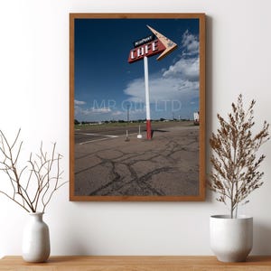 May include: A vintage roadside cafe sign with a red and white arrow pointing to the right. The sign reads "Midpoint Cafe" in white letters on a red background. The sign is on a white pole with a red stripe. The sign is in front of a blue sky with white clouds. The ground is cracked asphalt.