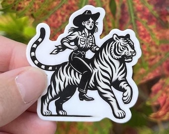 Op de afbeelding: Zwart-witte sticker van een vrouw met een cowboyhoed die op een tijger rijdt. De vrouw draagt een shirt met lange mouwen en een spijkerbroek. De tijger loopt met zijn bek open.
