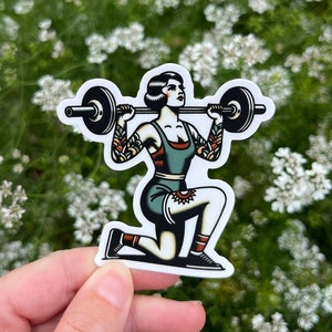 May include: A sticker featuring a vintage-style illustration of a woman lifting a barbell. She is wearing a green and red outfit and has a tattoo on her leg. The sticker is on a white background.