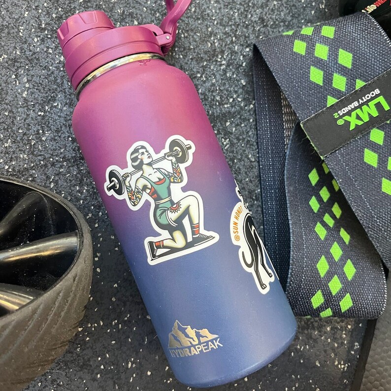 May include: A purple and blue stainless steel water bottle with a sticker of a woman lifting a barbell. The sticker says "Sun Hi Maui" and has a mountain range design. The bottle has the brand name "Hydrapeak" printed on it.