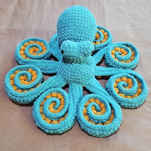 Puede incluir: Un pulpo de peluche hecho de hilo color verde azulado con una cabeza redondeada y ocho tentáculos rizados. Cada tentáculo presenta una parte inferior de color amarillo mostaza que crea un patrón en espiral. Textura tejida.