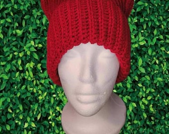 May include: A bright red, handmade crochet hat with cat ears. The hat is displayed on a white mannequin head against a backdrop of green foliage. The hat's texture is visible, showcasing the detailed crochet work.