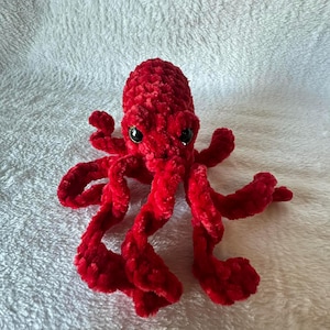 May include: A handmade, red crocheted octopus with black safety eyes. The plush toy features eight long, curly tentacles and a rounded head. The soft, textured yarn gives the octopus a cuddly appearance, perfect for a child's toy or nursery decor.