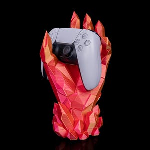 May include: A white and black game controller is held by a red and orange geometric hand-shaped stand. The stand has a crystal-like design. The background is black.