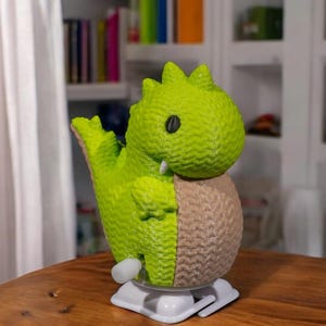 May include: A green and brown wind-up toy dinosaur with a cute face and a white base. The dinosaur is made of plastic and has a textured surface.
