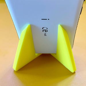 May include: A white smartphone is propped up on a yellow plastic stand. The stand has two triangular supports. The back of the phone has the text "ce mail" printed on it.