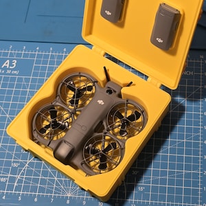 Puede incluir: Un estuche amarillo que contiene un dron gris con cuatro hélices y dos baterías grises. El dron tiene un diseño aerodinámico con protectores de hélice. El estuche está abierto, revelando el dron y las baterías en su interior.