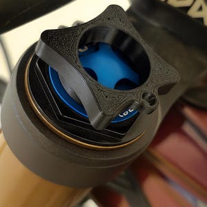 Puede incluir: Primer plano de un componente negro en forma de estrella con una superficie texturizada, posiblemente impreso en 3D, sobre una pieza de suspensión de bicicleta. El componente tiene una abertura central y se encuentra sobre una sección azul y dorada. La palabra "LOC" es visible.