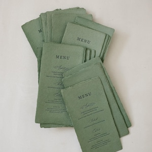 Może przedstawiać: Stos szałwiowych zielonych kart menu ze słowem "MENU" na górze. Karty mają teksturowany, ręcznie robiony wygląd papieru i zawierają elegancki skrypt dla przystawek, sałatek i dań głównych. Menu są ułożone w kształt wachlarza.