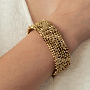 May include: A gold-colored mesh bracelet is worn on a person's wrist. The bracelet is wide and has a woven texture. The background is a white, textured fabric. The bracelet appears to be made of metal.