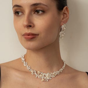 May include: A woman wearing a silver necklace and matching earrings. The necklace has a floral design with clear crystals and pearls. The earrings are a cascading design with crystals and pearls. The jewellery is set against a neutral background.