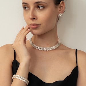 May include: A glamorous jewellery set comprising a double-strand necklace, matching bracelet, and stud earrings. The clear, round stones create a sparkling effect. The woman is wearing a black dress. The necklace measures approximately 40 cm.