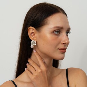 May include: A close-up of a pair of earrings. Each earring features a floral design with sparkling crystals and a large, dangling pearl. The earrings are set against a neutral background, highlighting their intricate details.