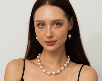 May include: A pearl necklace and matching earrings set. The necklace features large, round, off-white pearls strung together with gold-colored wire. The earrings have a single pearl suspended from a gold-colored hook. The jewelry is worn against a black top.