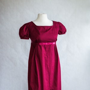 May include: A floor-length, deep magenta dress featuring a square neckline, short puffed sleeves with gathered detailing, and a high empire waistline accented by a thin, matching satin ribbon band that encircles the torso above the skirt.