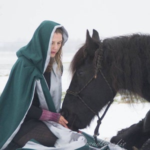 以下が含まれることがあります： 緑色のフード付きのマントを着た女性が、雪の中で黒い馬と一緒に座っています。馬は女性を見ていて、たてがみは編み込まれています。