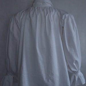 May include: A white long-sleeved shirt featuring a high collar, gathered fabric at the neckline, and sleeves that end in flared cuffs with tie closures. The garment is displayed on a mannequin, showcasing a loose fit and a solid white color.