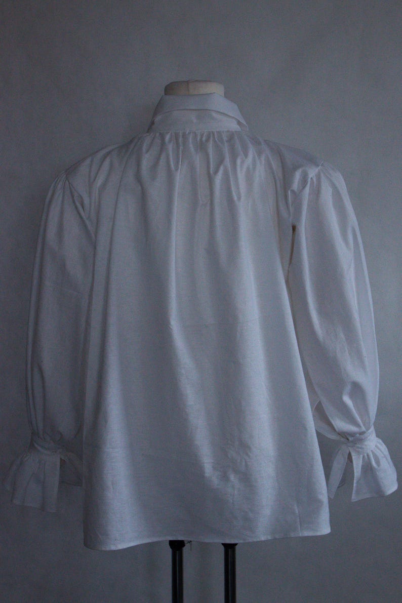 May include: A white long-sleeved shirt featuring a high collar, gathered fabric at the neckline, and sleeves that end in flared cuffs with tie closures. The garment is displayed on a mannequin, showcasing a loose fit and a solid white color.