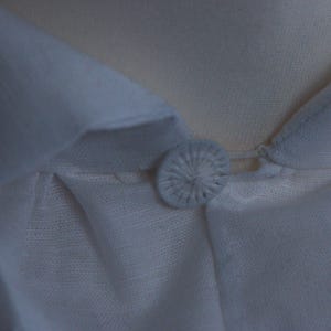 May include: A white shirt featuring a pointed collar and a single fabric-covered button at the neckline. The button is round with a radiating stitched pattern, securing the collar points together. The fabric has a visible, fine-textured weave.