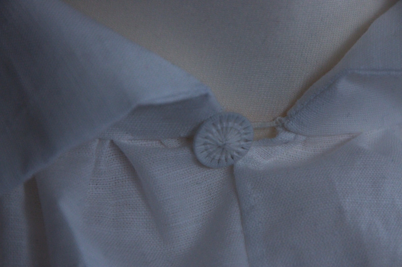 May include: A white shirt featuring a pointed collar and a single fabric-covered button at the neckline. The button is round with a radiating stitched pattern, securing the collar points together. The fabric has a visible, fine-textured weave.