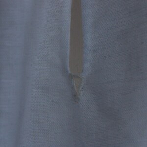 May include: A white fabric garment featuring a vertical slit opening at the neckline. The material has a visible woven texture with a slightly translucent quality. The edges of the slit are finished with stitching, showing a small triangular reinforcement.