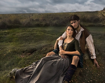 May include: A person wearing a dark green bodice with a lace-up front and a long grey skirt, sitting in front of another person wearing a light-colored long-sleeved shirt, a brown vest, and a dark kilt with blue accents in a grassy field.