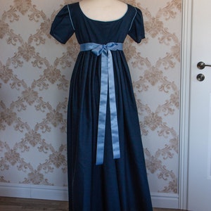 May include: A floor-length dark blue dress with short sleeves and a scoop neckline. The garment features light blue piping along the neckline and sleeve edges, and a light blue satin ribbon tied in a bow at the high waistline.