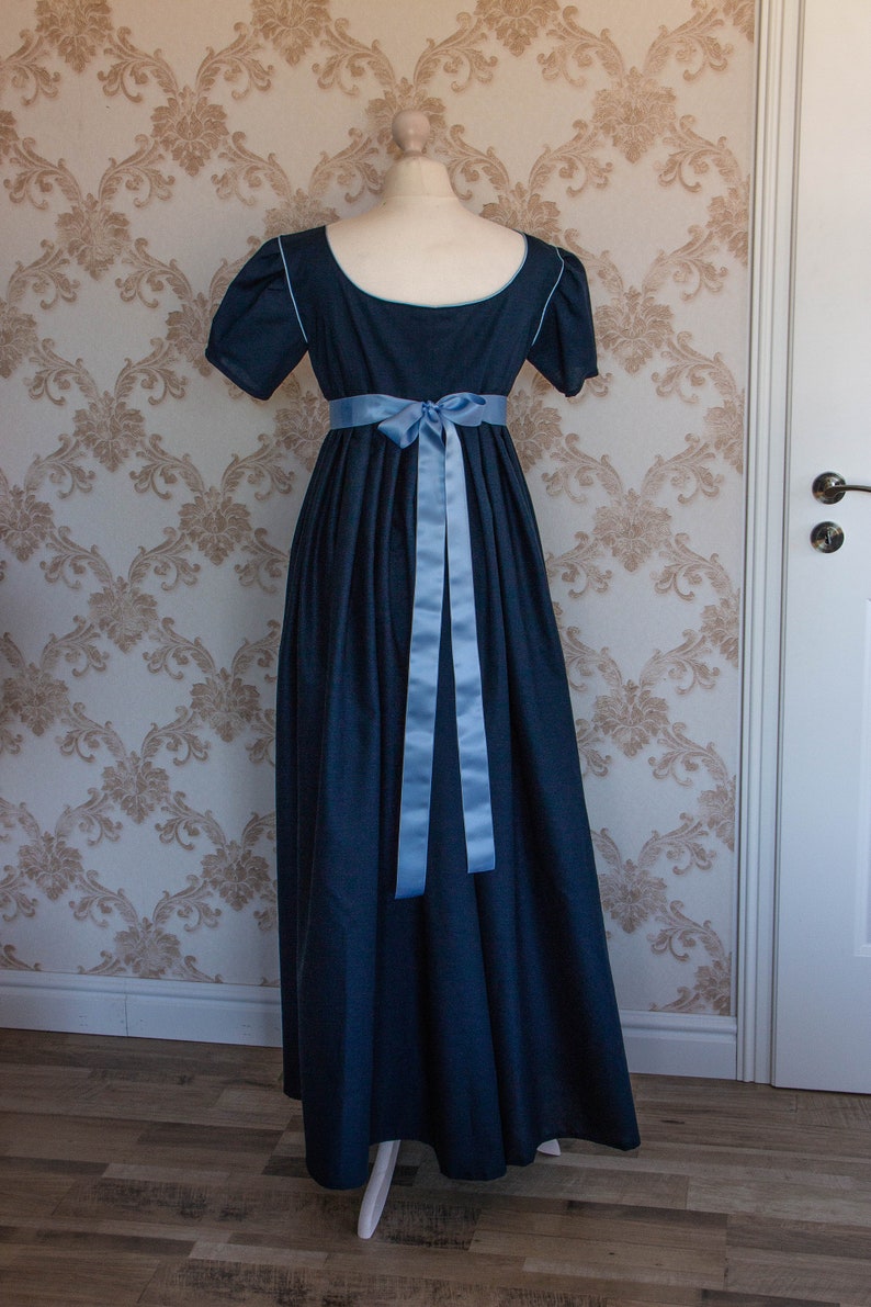 May include: A floor-length dark blue dress with short sleeves and a scoop neckline. The garment features light blue piping along the neckline and sleeve edges, and a light blue satin ribbon tied in a bow at the high waistline.