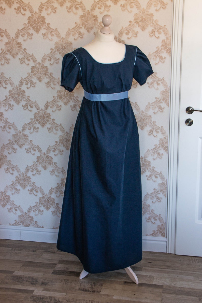 May include: A floor-length dark navy blue dress featuring short puff sleeves, a square neckline with light blue piping, and a wide light blue ribbon sash positioned at the empire waistline. The garment is displayed on a mannequin form.