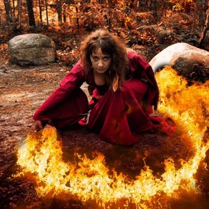 May include: A person with wavy brown hair wearing a long-sleeved, floor-length deep red dress with a wide skirt. The person is crouching on the ground, holding a curved blade that creates a bright, swirling arc of orange and yellow flames.